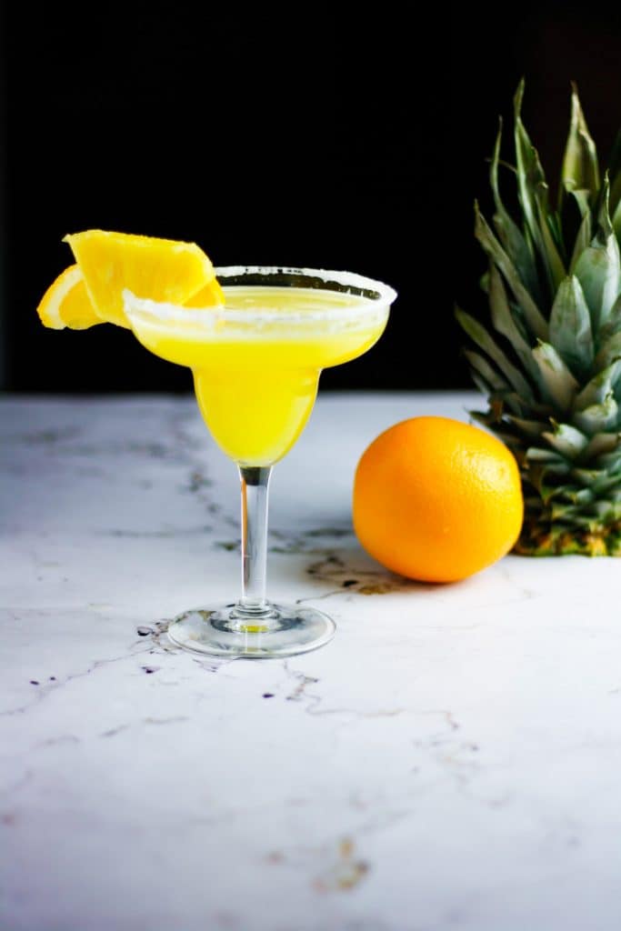 Glass of pineapple orange margarita on a table with an orange and pineapple in the background