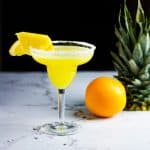 Glass of pineapple orange margarita on a table with an orange and pineapple in the background