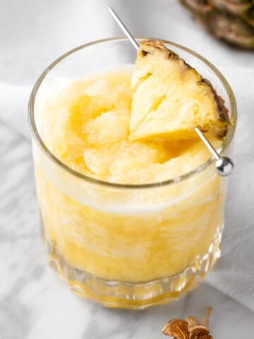 A pineapple slushie garnished with a pineapple slice on a white marble table.