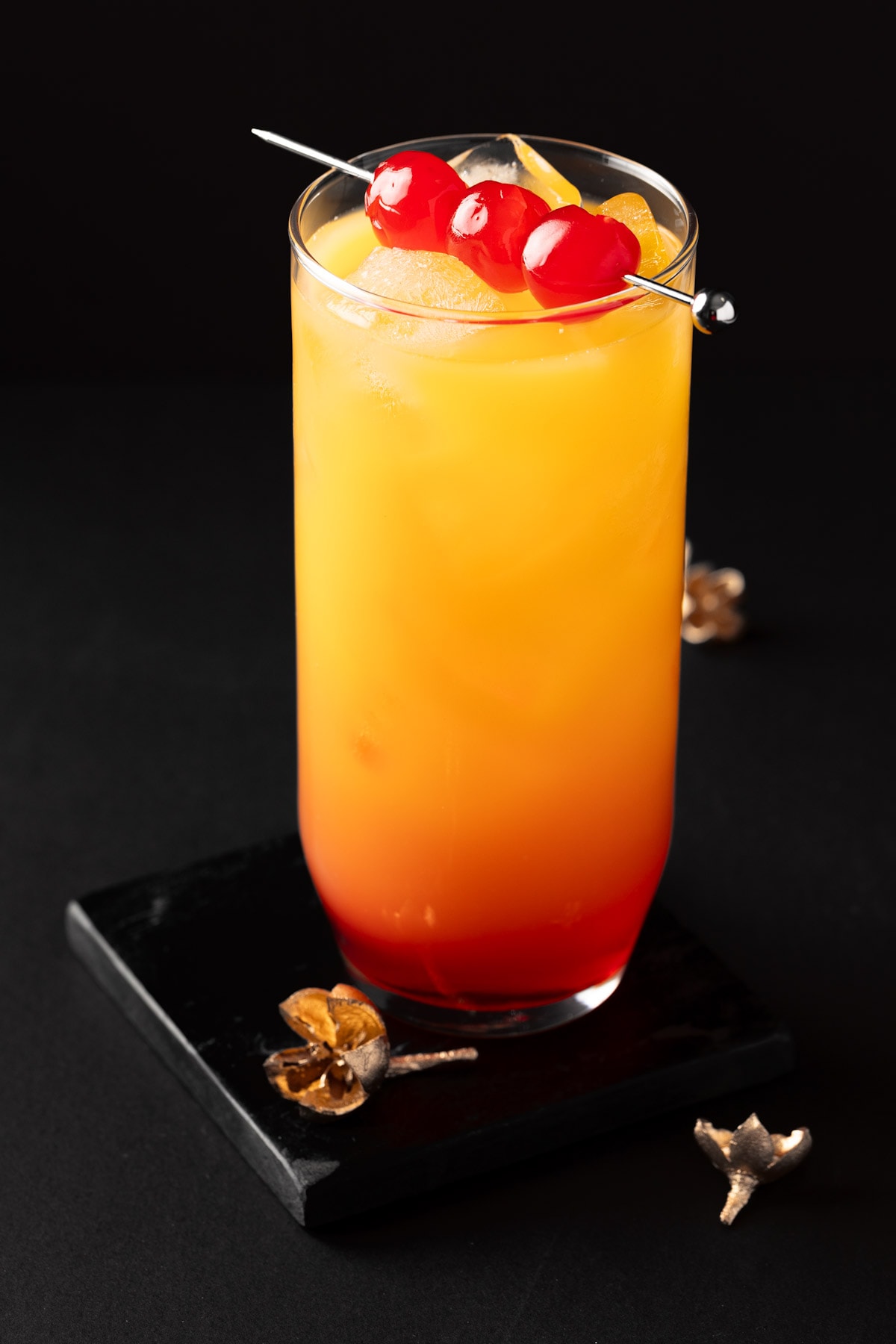 An yellow and red Tequila Sunrise Cocktail with pineapple juice, on a black coaster with a black background.