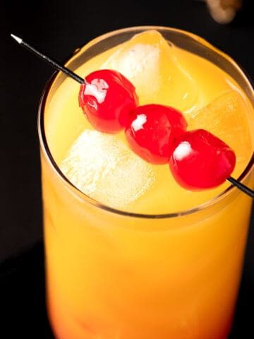 A Pineapple Tequila Sunrise garnished with maraschino cherries on a black background.