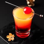 An Upside Down Pineapple Cake Shot garnished with a maraschino cherry on a black background.