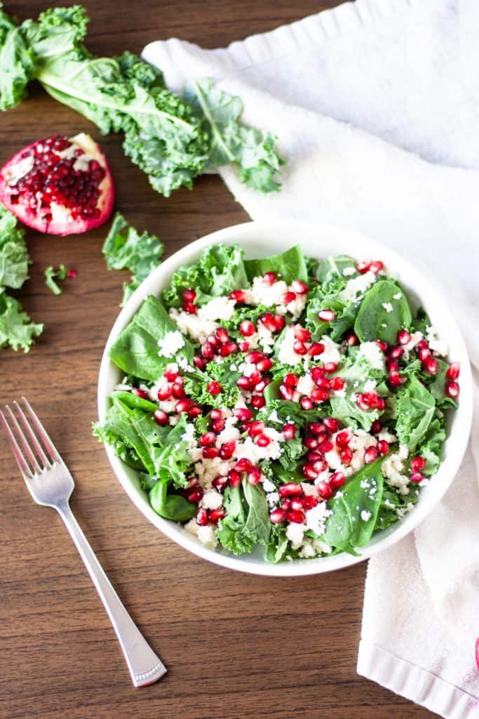 Overhead view of a bowl of pomegranate kale and feta salad on a wooden table with a fork beside it