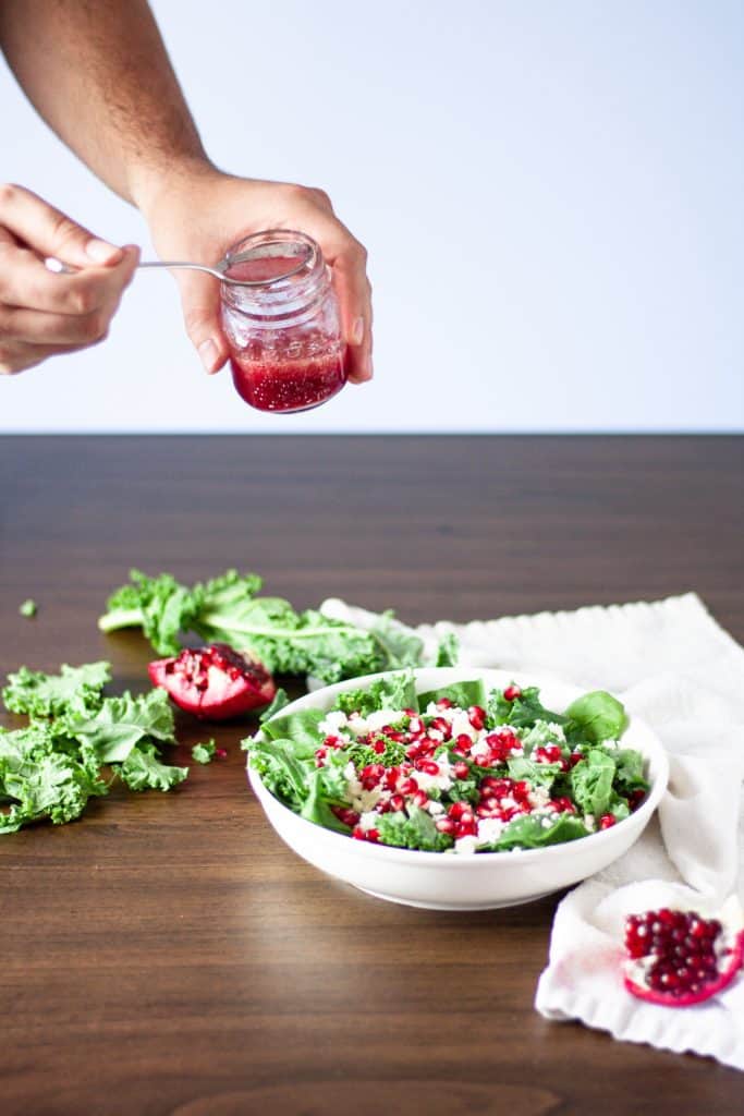 Action shot of a man’s hands holding the jar of pomegranate vinaigrette and drizzling it over the salad