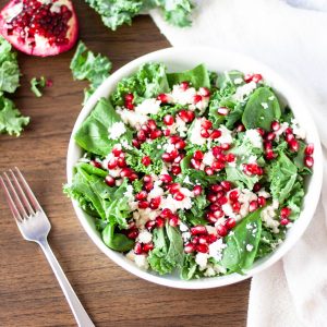 Overhead view of a bowl of pomegranate kale and feta salad on a wooden table with a fork beside it