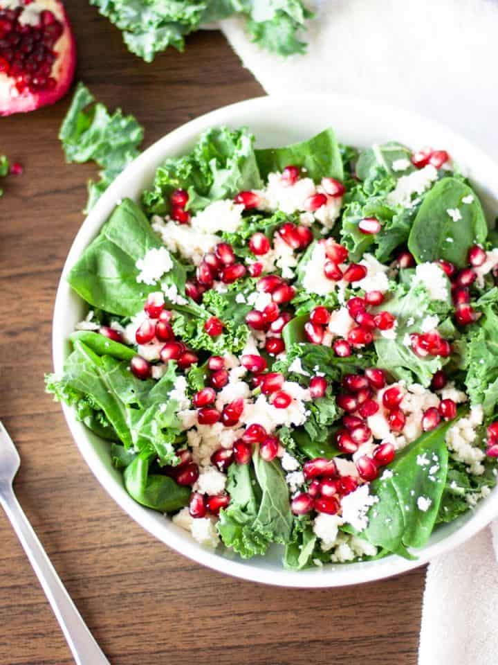 Overhead view of a bowl of pomegranate kale and feta salad on a wooden table with a fork beside it
