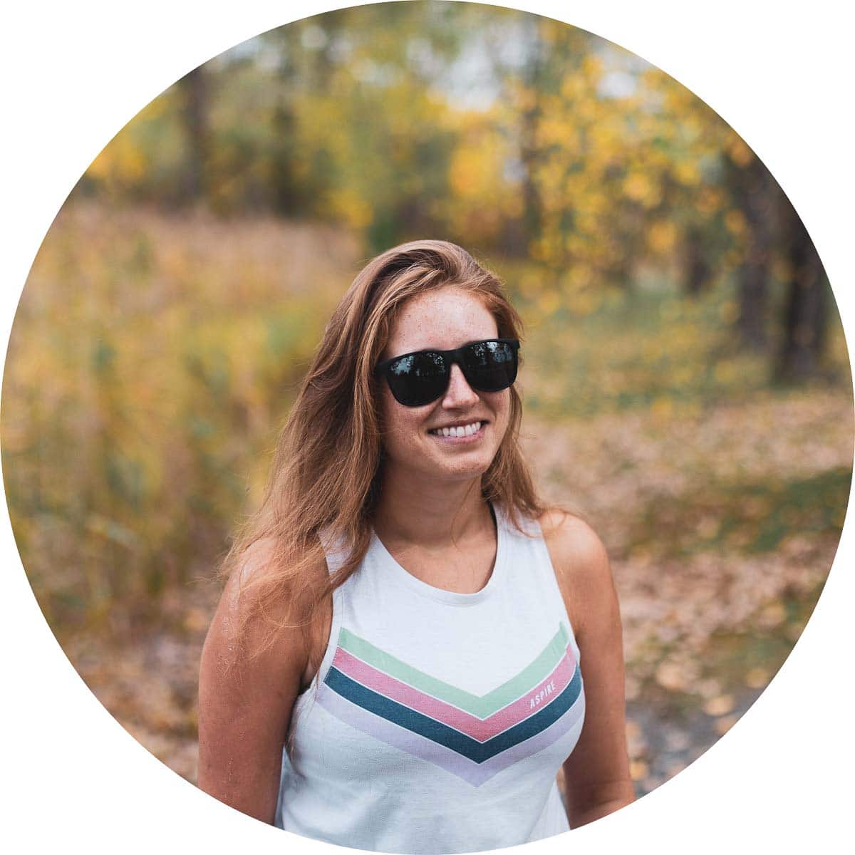 Ali in a white tank top wearing sunglasses, with trees with yellow leaves in the background.