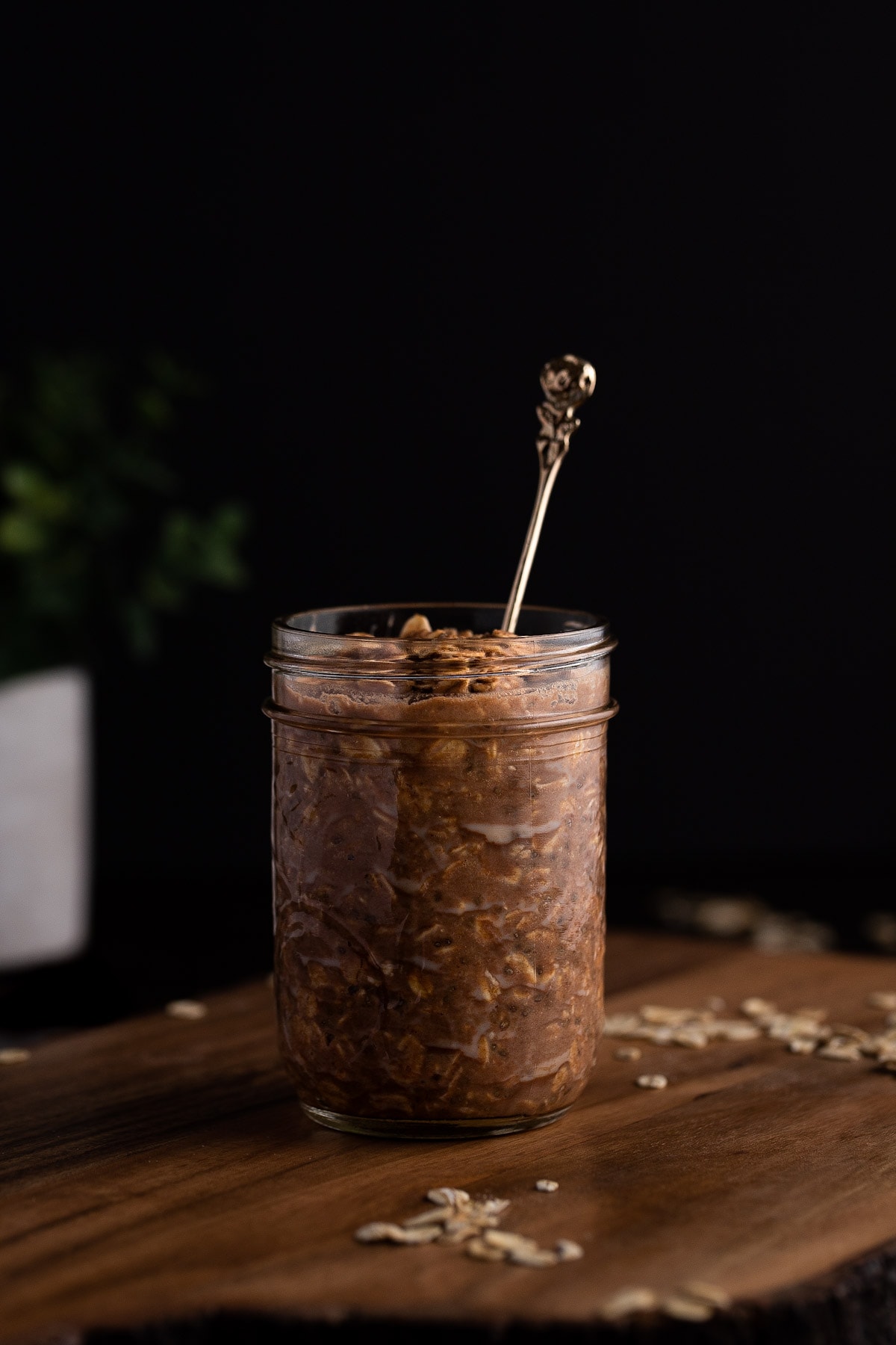 A jar of protein overnight oats on a wooden board with oats scattered at the base, on a black background.