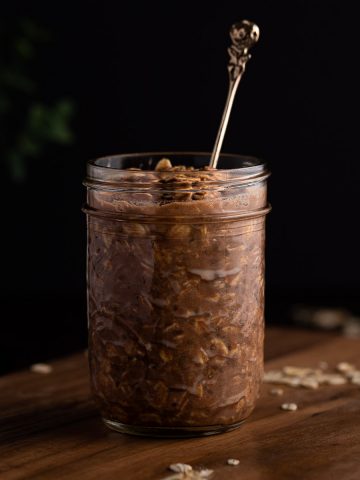 A jar of protein overnight oats on a wooden board with oats scattered at the base, on a black background.
