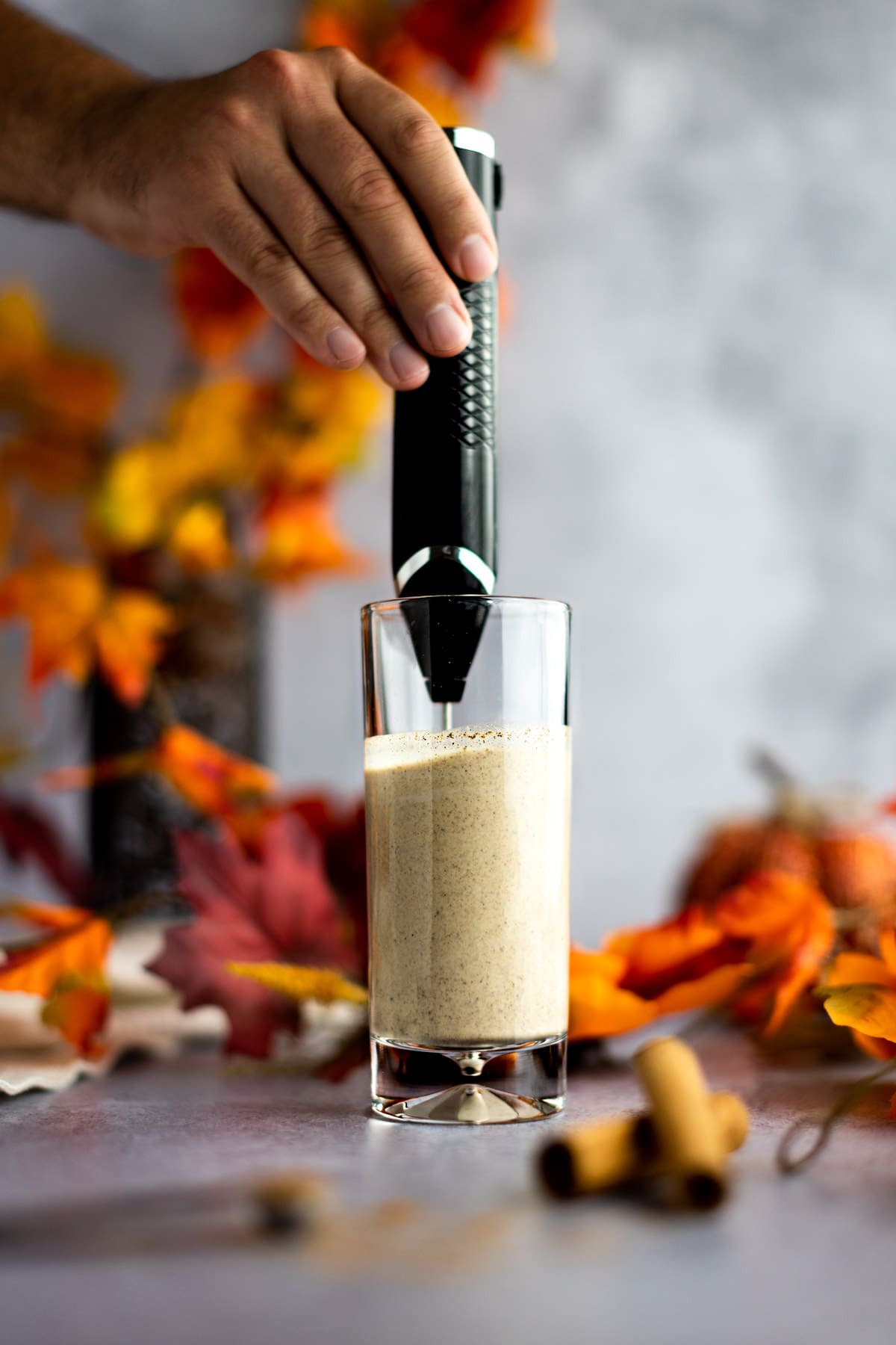 Hand holding a frother, blending the pumpkin foam in a tall, clear glass, with leaves in the background.
