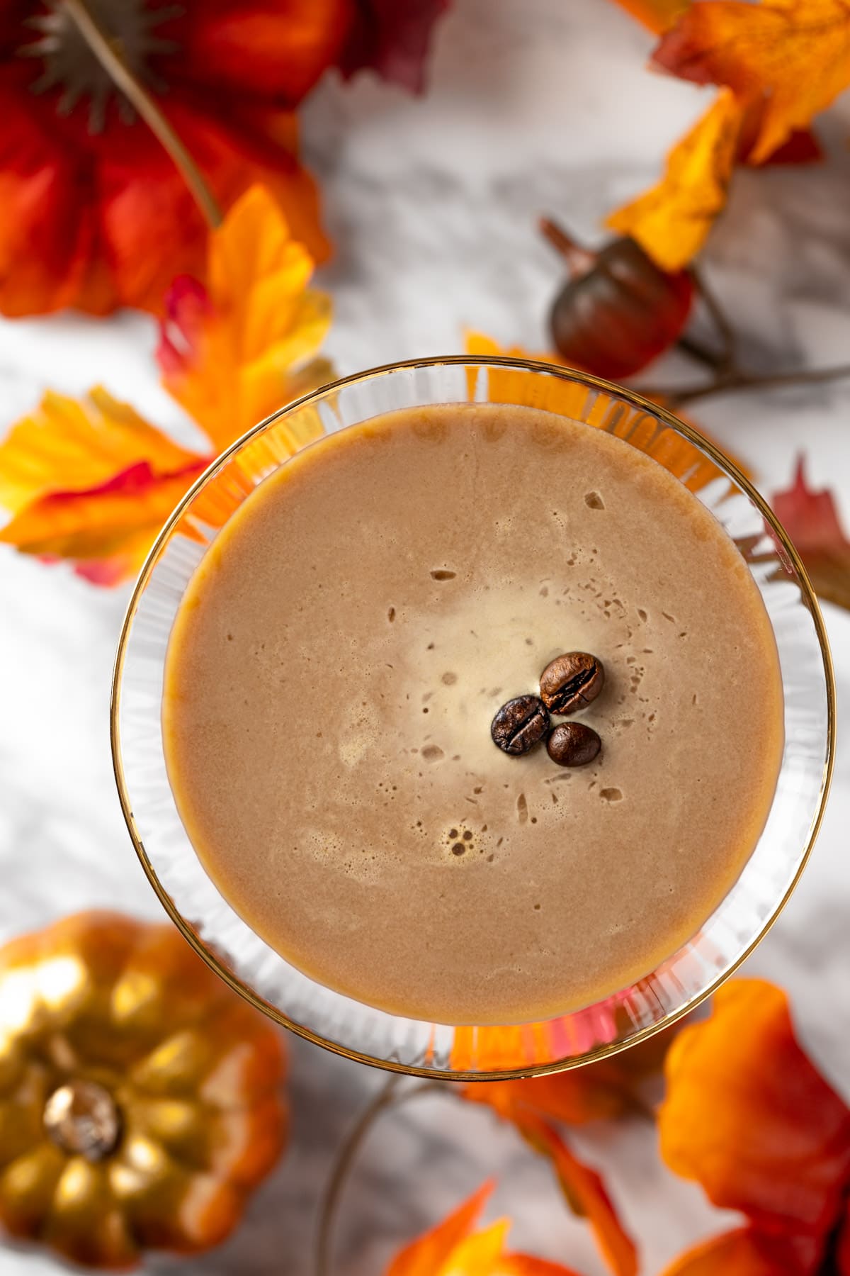 Overhead view of a pumpkin spice espresso martini on a white marble table next to pumpkins and leaves.