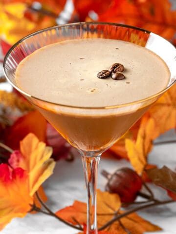 A pumpkin spice espresso martini garnished with three coffee beans, with fall leaves in the background.