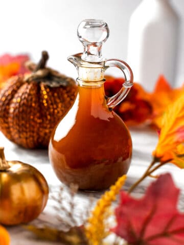 A bottle of pumpkin spice syrup surrounded by fall leaves and pumpkins.