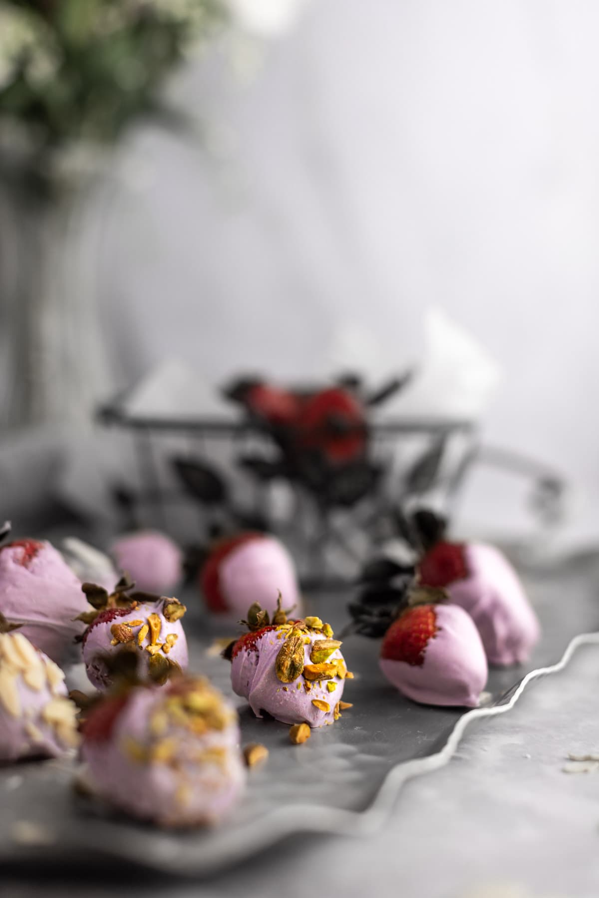 A tray of purple chocolate covers strawberries, topped with crushed pistachios and almonds, with a glass vase of flowers in the background.
