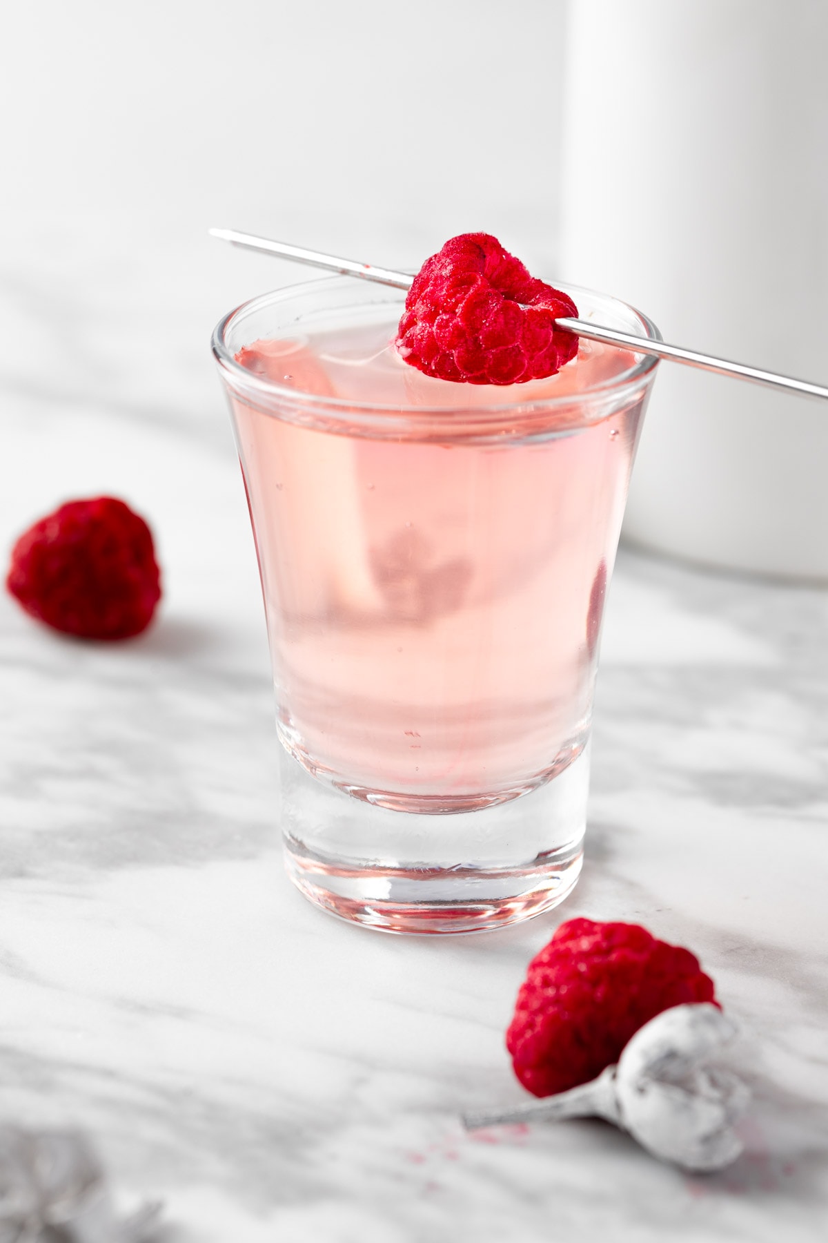 A Raspberry Lemon Drop Shot garnished with a fresh raspberry, on a white table.