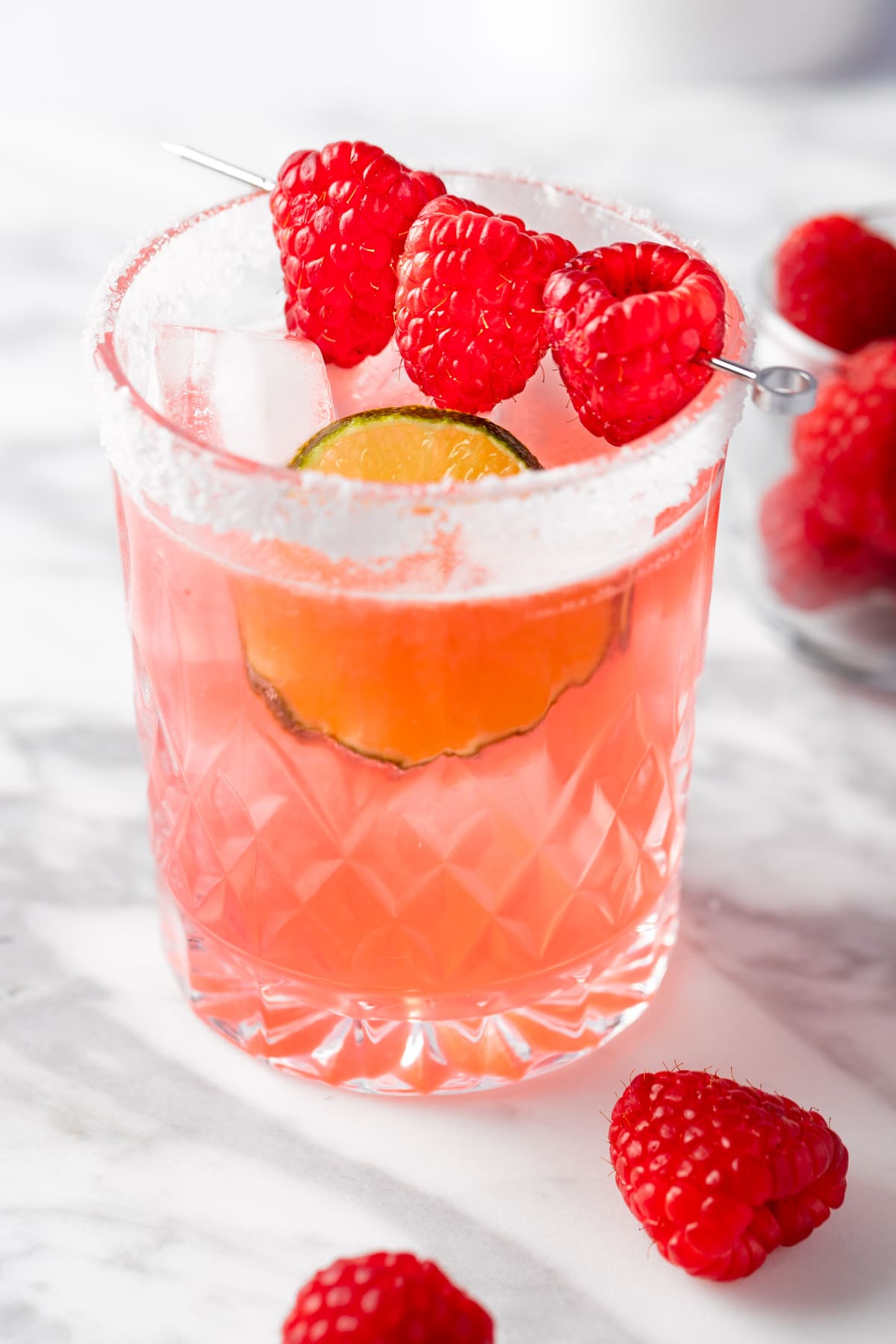 A raspberry margarita garnished with fresh raspberries, sitting on a white marble table.