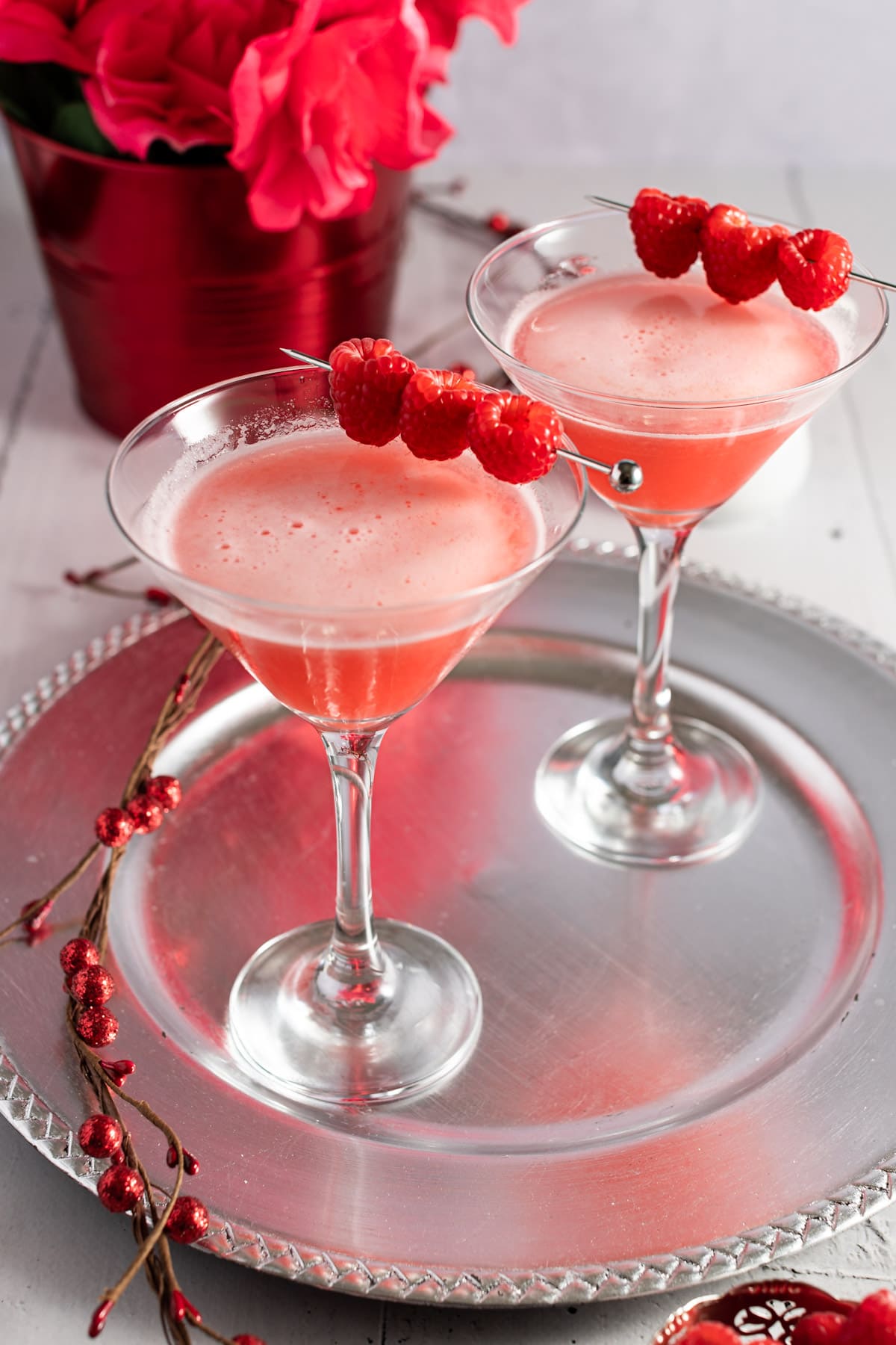 Two martinis on a silver plate, garnished with fresh raspberries, with a red bucket with red roses in the background.