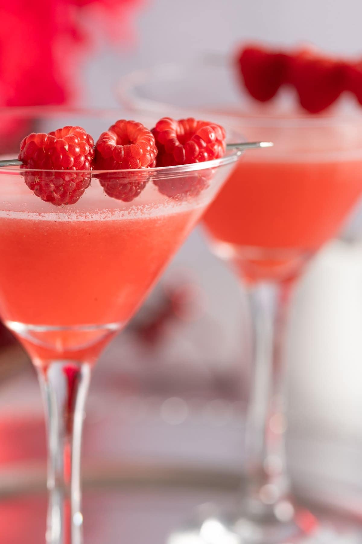 Up close view of the edge of a martini glass, garnished with raspberries and another martini in the background, slightly out of focus.