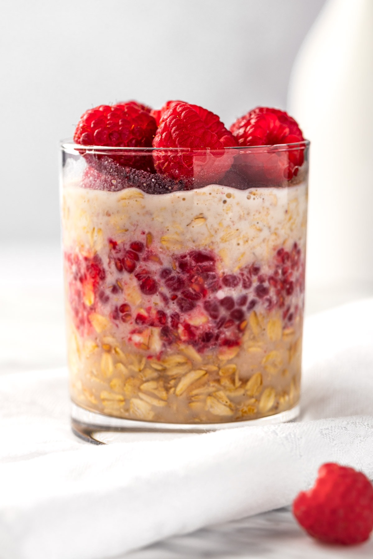 A glass of raspberry overnight oats, topped with fresh raspberries, sitting on a white napkin.