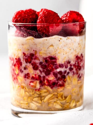 A glass of raspberry overnight oats, topped with fresh raspberries, sitting on a white napkin.