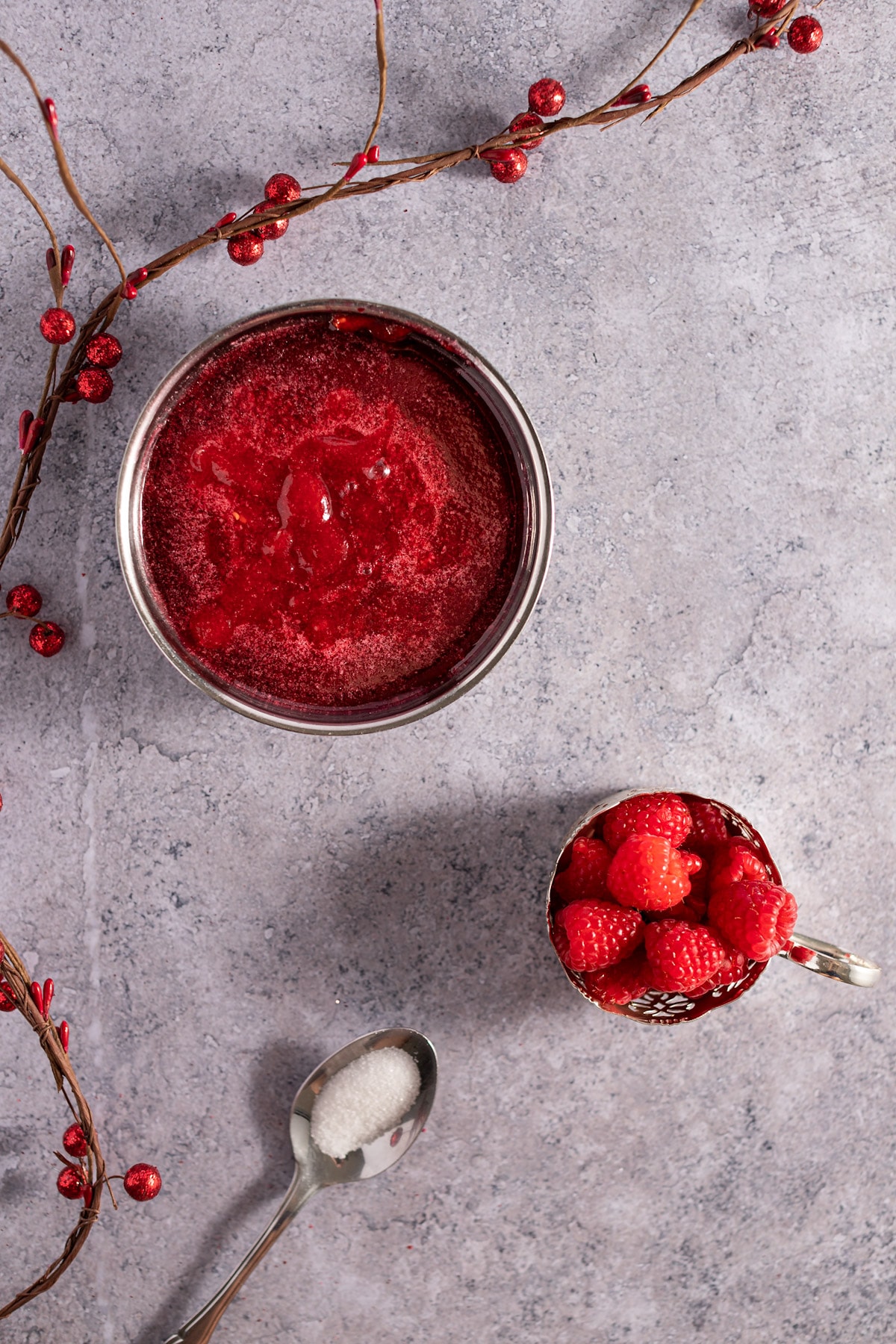 Overhead photo of raspberry syrup in a metal bowl, with a container of fresh raspberries, spoonful of sugar and garland of red berries.