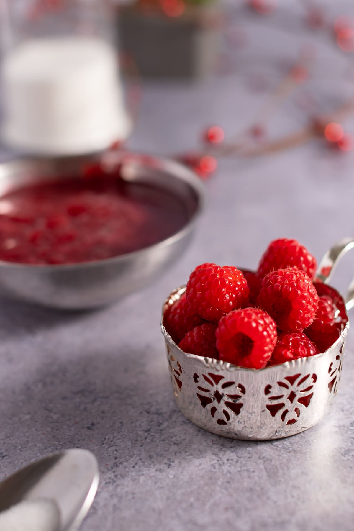 A small silver container filled with fresh raspberries sitting on a grey tabletop, with syrup and a garland of red berries in the background.