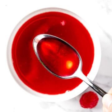 Overhead view of a spoonful of bright red raspberry syrup, with a white marble background.