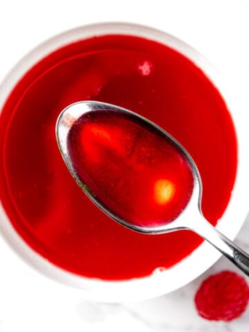 Overhead view of a spoonful of bright red raspberry syrup, with a white marble background.