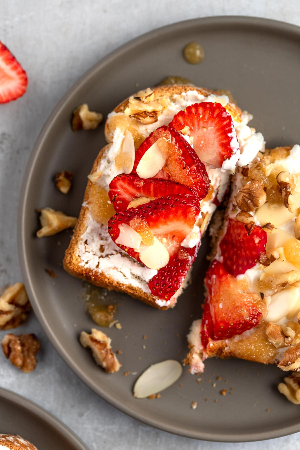 Ricotta honey toast topped with walnuts, almonds and sliced strawberries on a grey plate.