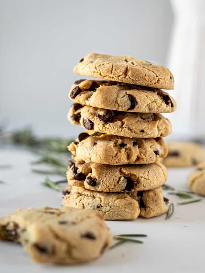 A stack of rosemary chocolate chip cookies with a half eaten cookie in the foreground and a white vase in the background