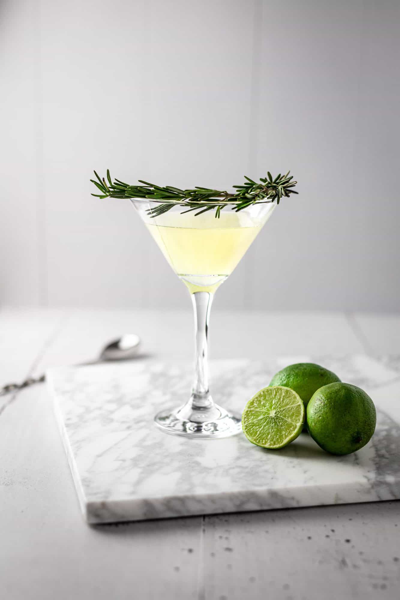 Rosemary gimlet with a branch of fresh rosemary as garnish around the glass, three lines sitting on the marble table beside the cocktail.