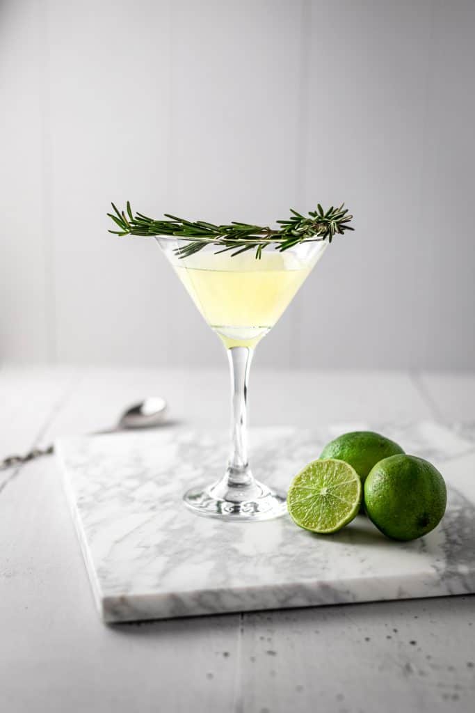 Rosemary gimlet with a branch of fresh rosemary as garnish around the glass, three lines sitting on the marble table beside the cocktail