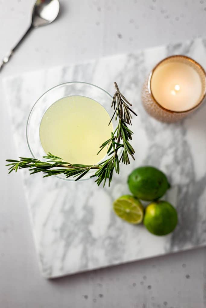 Overhead view of gimlet with rosemary garnish, 3 limes and a lit candle on a white marble board