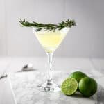 Rosemary gimlet with a branch of fresh rosemary as garnish around the glass, three lines sitting on the marble table beside the cocktail