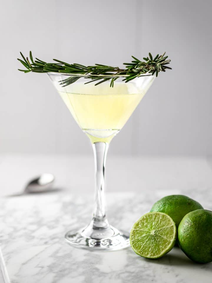 Rosemary gimlet with a branch of fresh rosemary as garnish around the glass, three lines sitting on the marble table beside the cocktail