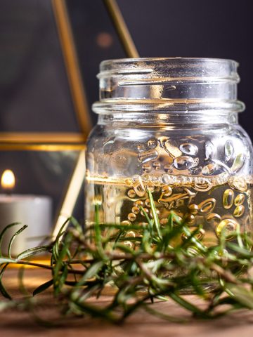 A small glass jar filled with rosemary syrup, with fresh rosemary in front and a candle in the background.