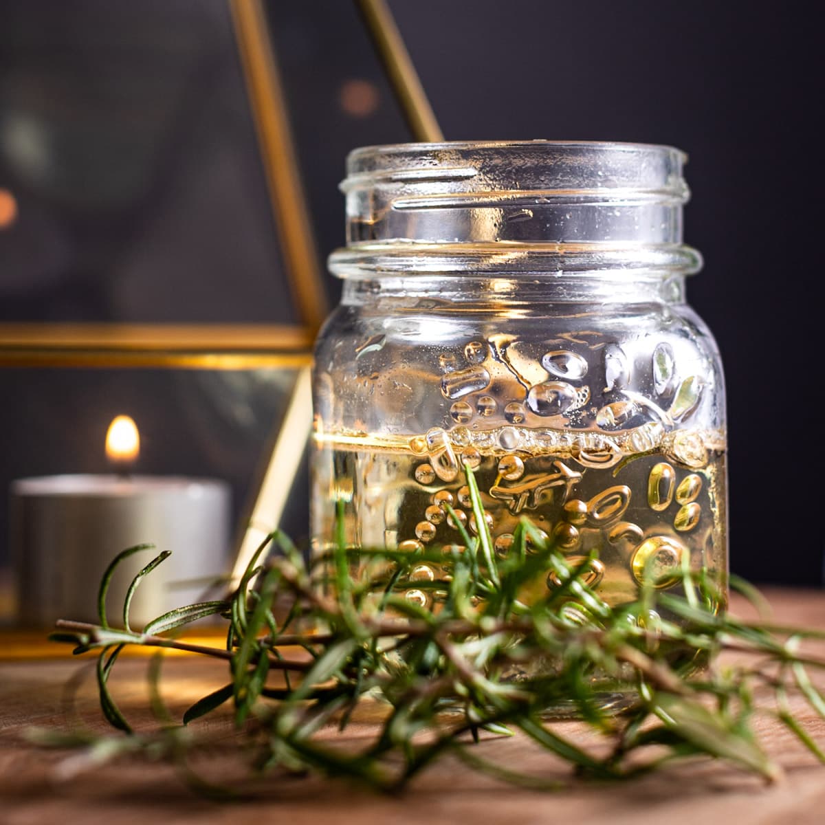 Glass jar of rosemary simple syrup, with fresh rosemary in front, on a black background. 