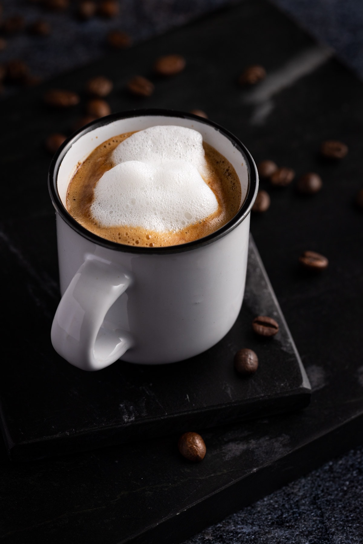Doppio macchiato in a small white cup on a black coaster with coffee beans scattered on the table.