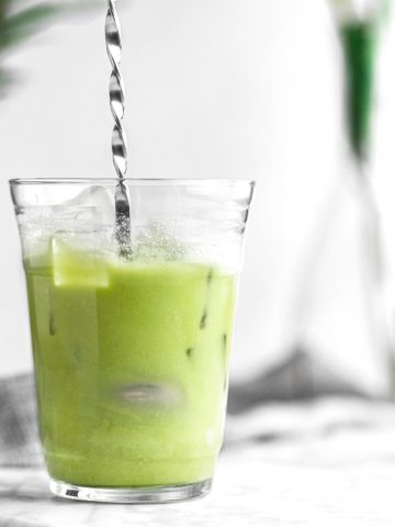 Glass of green starbucks pineapple matcha being stirred with a long metal spoon, on a grey background.