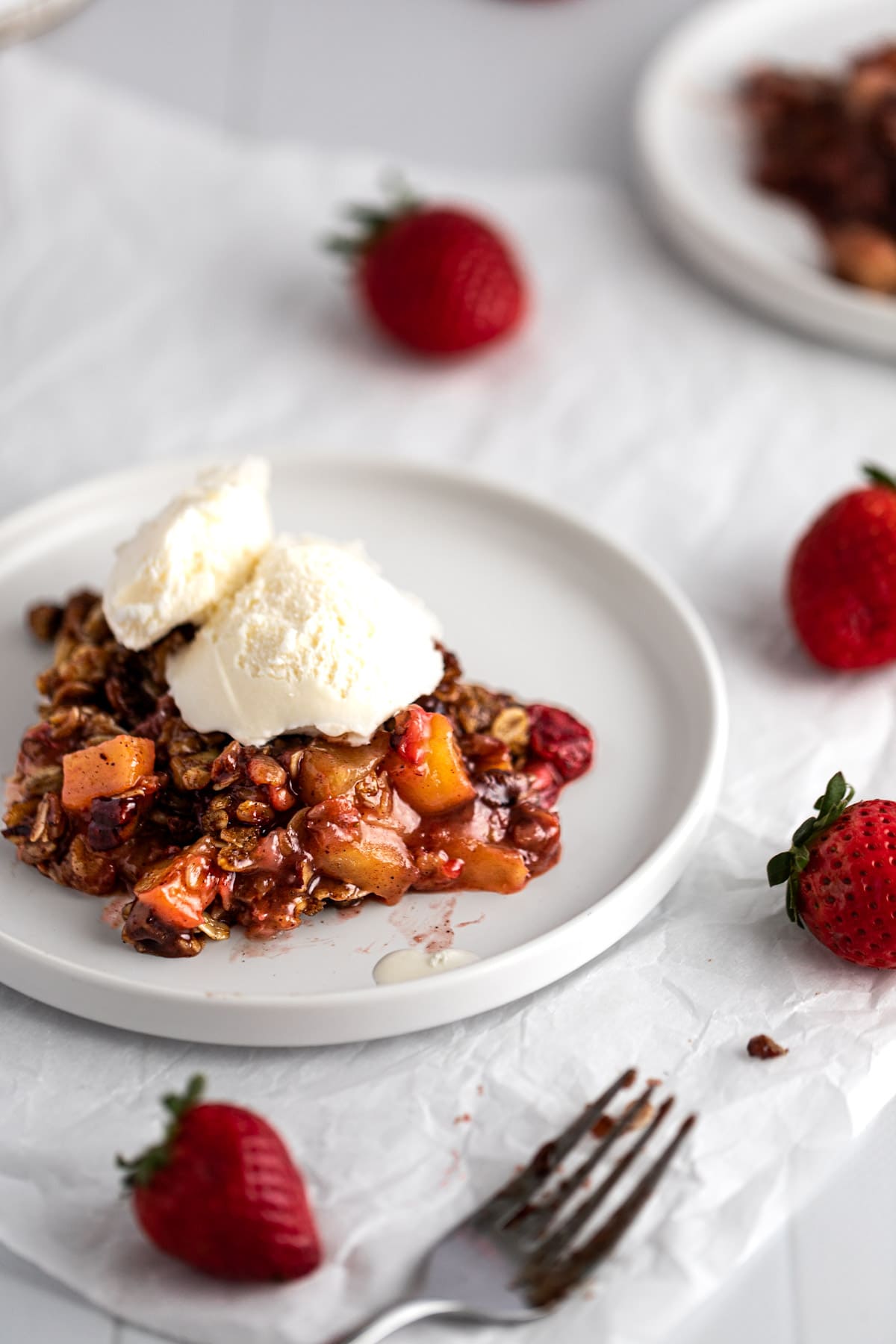 A piece of strawberry apple crisp topped with a scoop of vanilla ice cream, on a white round plate.