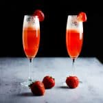 Two flutes side by side filled with red strawberry bellini and strawberries on the table