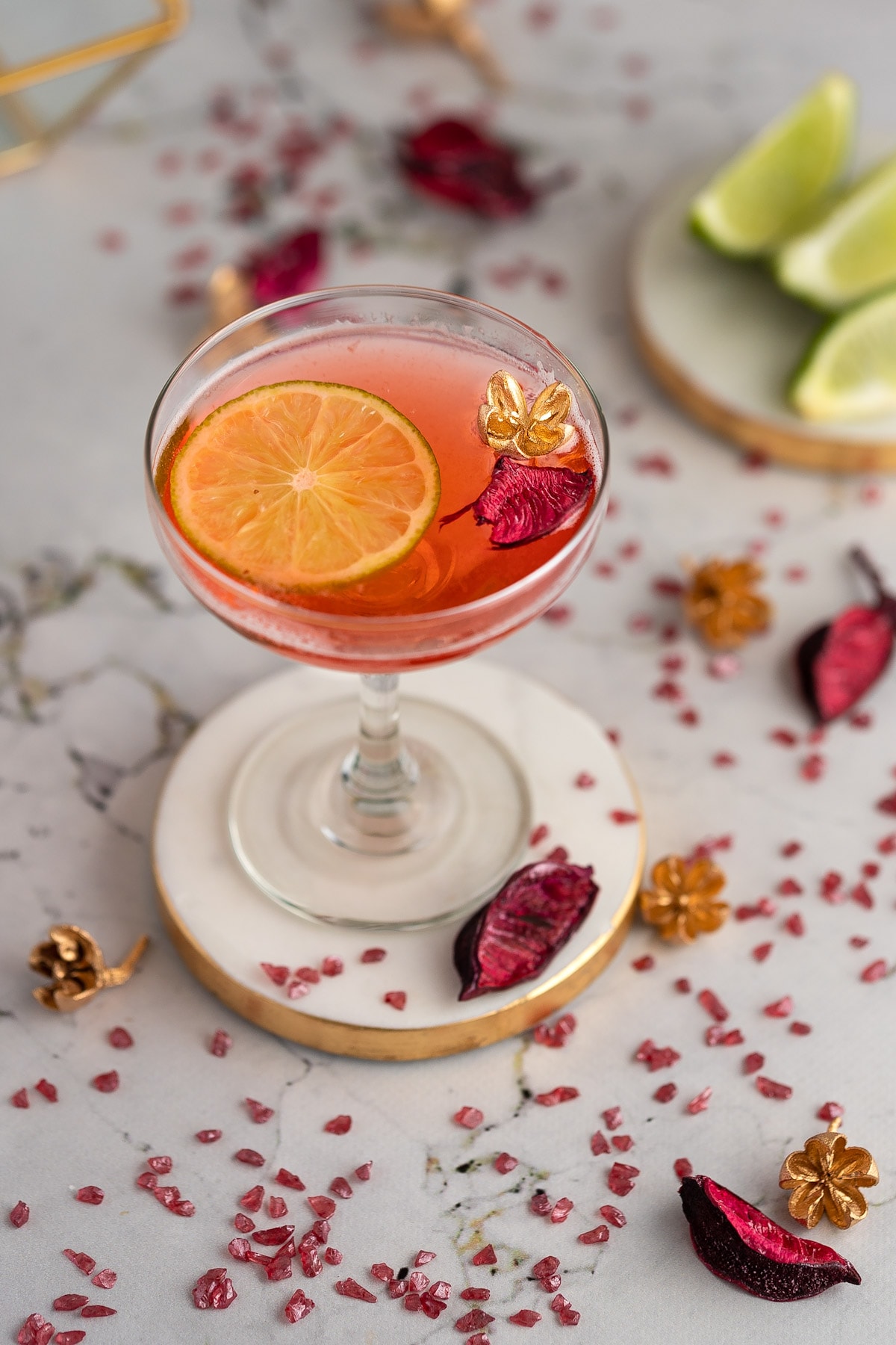 Strawberry gimlet on a white and gold coaster, on a marble table, with pink and gold flowers scattered around and sliced limes in the background.