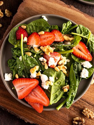 Strawberry goat cheese salad with spinach, walnuts and pine nuts on a dark grey plate on a wooden serving board.