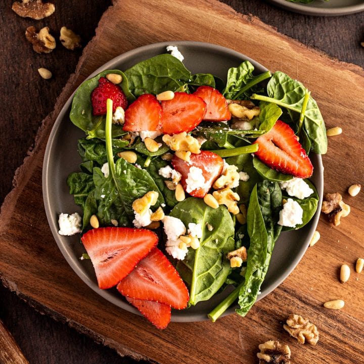 Strawberry goat cheese salad with spinach, walnuts and pine nuts on a dark grey plate on a wooden serving board.