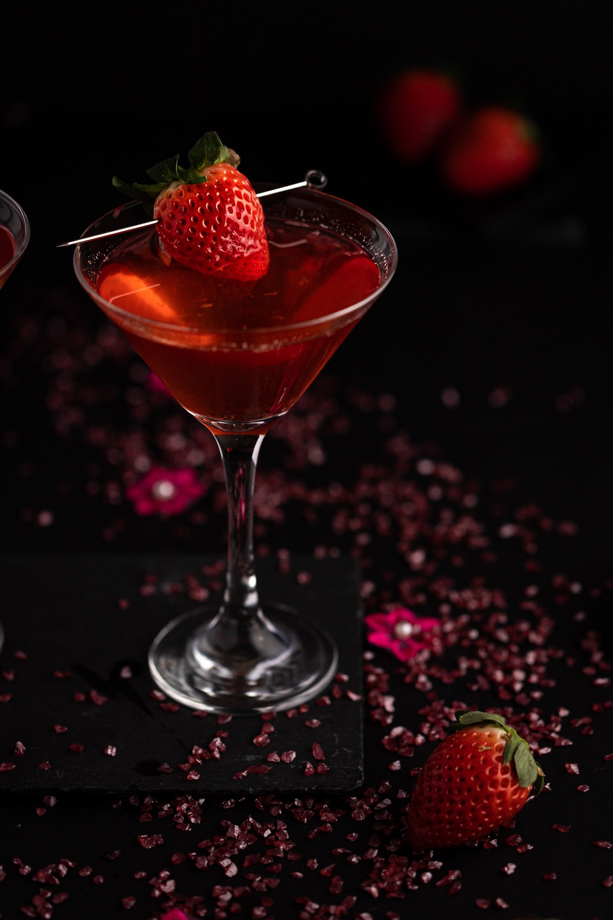 Strawberry martini, garnished with a strawberry, on a black background, with pink pebbles and flowers scattered around.