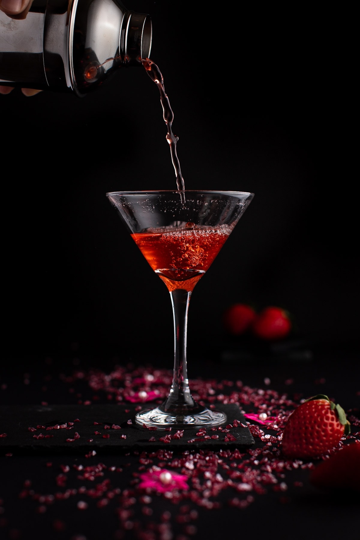 Strawberry martini being poured from a cocktail shaker into a martini glass, on a black background.
