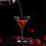 Strawberry martini being poured from a cocktail shaker into a martini glass, on a black background.