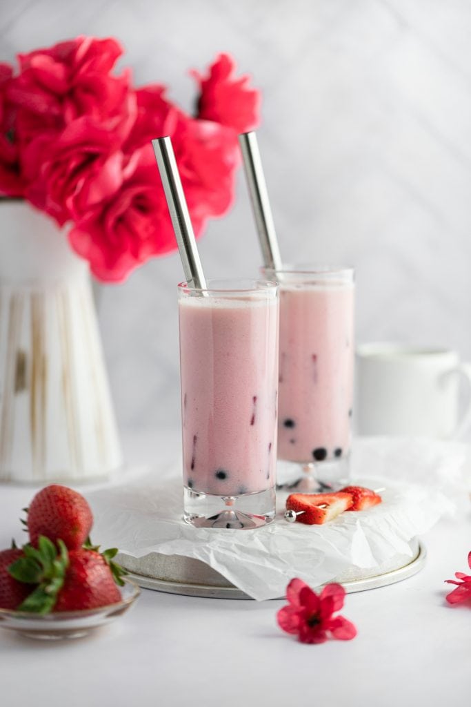 Two tall glasses of strawberry milk tea with a vase of red roses in the background and a plate of strawberries in the foreground.