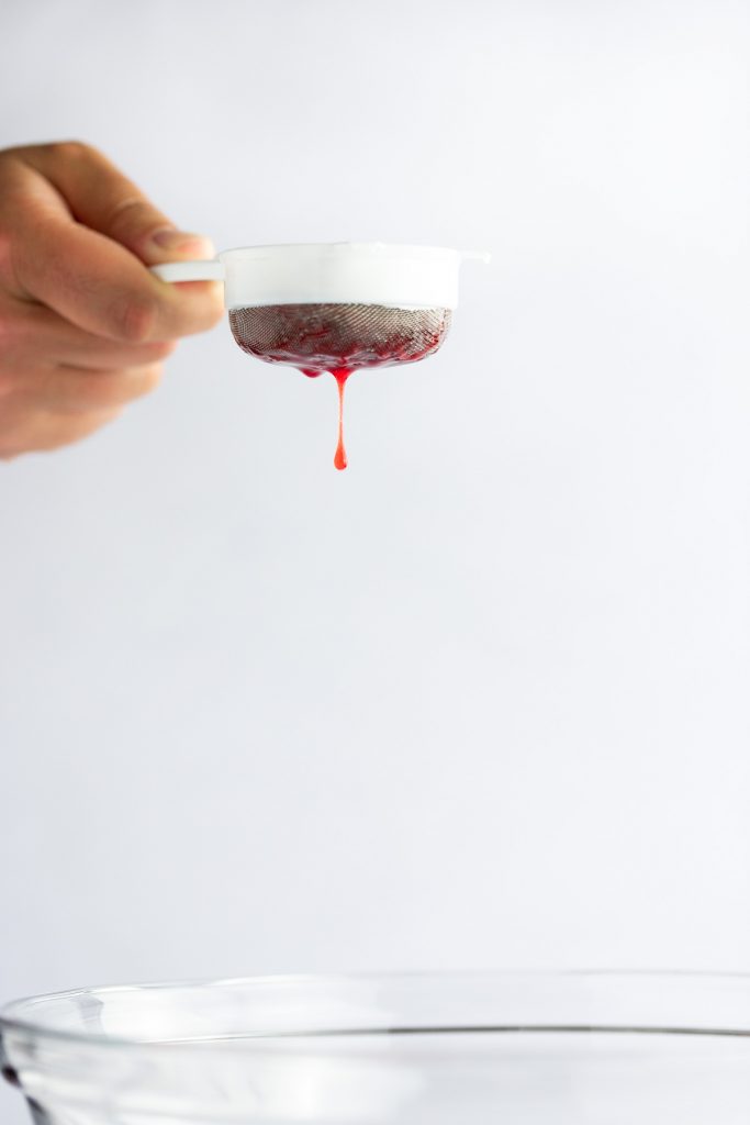 A hand holding a small fine mesh sieve draining the strawberry syrup, with a drip falling into a bowl underneath.