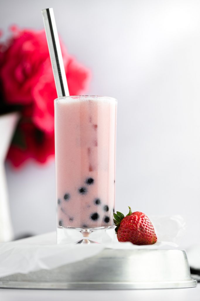 A close up view of a tall glass of strawberry bubble tea with a metal straw, with a single strawberry sitting next to the glass.